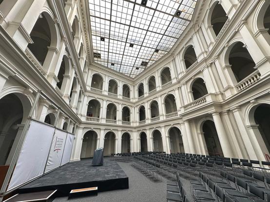 Ein Innenbereich mit einer großen, gewölbten Architektur mit weißen Wänden und mehreren Ebenen. Der Bereich ist mit Reihen grauer Stühle ausgestattet, die einem Podium auf einer schwarzen Bühne zugewandt sind. Eine Glasdecke lässt natürliches Licht die Umgebung beleuchten und trägt zum geräumigen und hellen Raum bei.