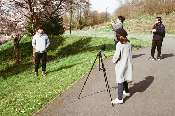 This image captures a serene outdoor scene in a park. Four individuals are present, engaged in what seems to be a casual photoshoot. On the left, a man stands near a blooming tree, its branches adorned with white flowers. He is dressed in a light jacket and dark pants, smiling gently. In the center, a camera is mounted on a tripod, aimed towards the group. A woman in a long coat stands beside the camera, observing the scene. Behind the camera, two more individuals, one holding a phone and the other standing casually, complete the group. The park is lush with green grass and trees, and the path they stand on is paved and clean.