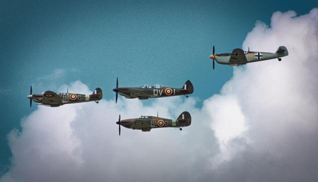 Four vintage military aircraft are flying in formation against a backdrop of a clear blue sky with scattered clouds. Three of the planes feature British insignia (two Spitfires and a Hurricane) and the fourth (a Messerschmitt) displays German insignia.