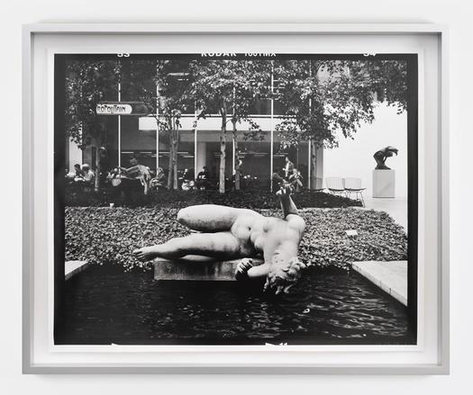 Image: Gerard Byrne, M7 / Roll 32 / Exp 08, 2008 – ongoing, hand-printed silver gelatin print, edition of 3, 38.6 x 47.8 cm print size | black-and-white photo taken in the courtyard of, possibly, of a museum of modern art, probably in a French-speaking area, judging by a sign that we can see with letters ‘opolitain’; there is a modern-style in the background, a modern-style sculpture on the right side of the image, what appears to be a café area in front of the building, with some narrow tree trunks and leaves aboe, then foliage as ground cover closer to the camera, then a pool of dark water with a pale statue in classical style, probably on its side, of a curvy naked figure with upraised harms as though fearing a fall into the water.• “Kerlin Gallery is pleased to present The Struggle with the Angel, a new exhibition by Gerard Byrne.    In Autumn 2008, Gerard Byrne bought a used camera from a friend for cash. For 17 years since that day, the camera (a Mamiya 7 analogue stills…”
