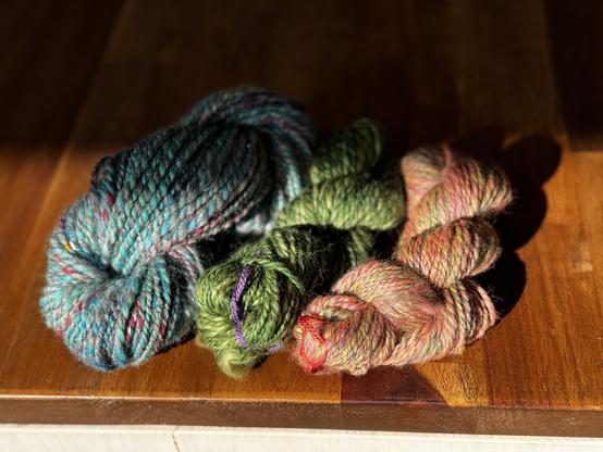 Three small skeins of colourful handspun 2ply wool sit in a narrow sliver of sun on a kitchen bench. Photos can’t bring out the sparkle of the larger blue and grey skein that is flecked with reds and yellows. The middle skein is a mix of greens from yellowish to forest, and the right skein is a colourful blend of yellows, pinks and greens.