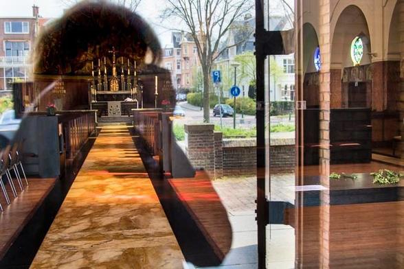 I took a selfie here in (and through) the glass interior doors of the Antonius Abtkerk in The Hague. It's a strange photo because you only see the interior of the church where my image blocks the daylight. Around it, you see the other side of the street reflected in normal daylight. On the left halve of the photo you see within the contours of my image (only upper body) a long, vertical, marble, orange-yellow illuminated path, wide at the bottom of the photo due to the perspective and narrow in the distance. At the end of the path, at the height of my face and camera, is a large golden cross with behind it high, golden (by the light) pipes of a church organ. So you see in my contours exactly what I see through the viewfinder of the camera.