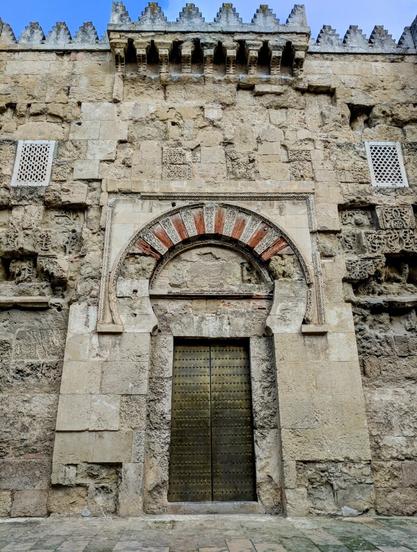 A weathered stone facade, likely part of a historic building such as the Mezquita-Catedral de Córdoba, dominates the frame. The stonework is a light beige, showing significant signs of age and erosion, with varied textures and uneven surfaces.
A prominent horseshoe archway, characteristic of Moorish architecture, frames a set of aged, dark metal double doors. The arch is outlined in a lighter stone and features alternating red and white voussoirs. Above the archway, there is a rectangular panel with intricate, though worn, carvings.
Flanking the central archway are sections of the stone wall with further decorative elements, including what appear to be blocked-up windows or niches with geometric patterns. The top of the facade is crenellated, with a series of rectangular merlons against a partly cloudy blue sky.
The ground in front of the facade is paved with light-coloured stone slabs, showing some signs of wear.