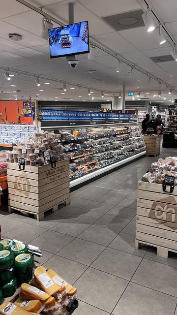 The image features the interior of a supermarket, showcasing various food items. On one side, there are shelves displaying packaged foods and baked goods, while the foreground includes wooden crates with snacks and cheese. A security monitor is visible hanging from the ceiling.