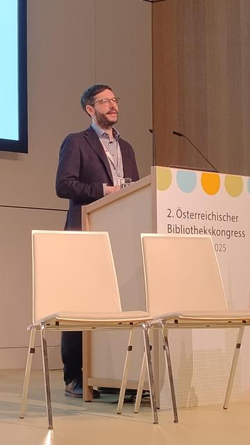 A close-up of Peter Kraker at the podium during the 2nd Austrian Library Congress. He is mid-speech, wearing a blazer, glasses, and a lanyard, standing behind a podium branded with the event's colourful logo and title.