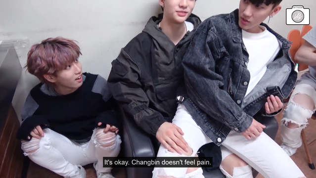 Changbin sits on Hyunjin's lap with his legs spread. Hyunjin has just slapped his thigh through a rip in his jeans. Han crouches on the floor next to them and looks surprised and/or impressed. Caption: "It's okay. Changbin doesn't feel pain."