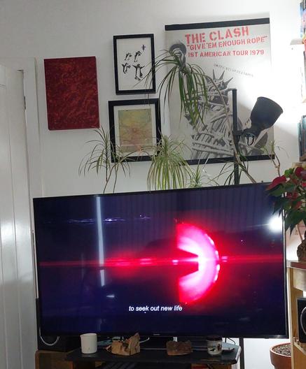 A large TV with the opening credits to Star Trek Strange New Worlds on it. There is a tall lamp, a large plant, and a plain white wall with art on it behind the TV. The stand it is on also has pretty bits of wood and a couple of candles on it.