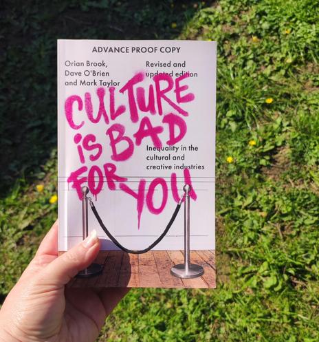 A white woman's hand holding a book in the sunshine, against a backdrop of grass and little yellow flowers. 

The book cover is of a white wall, a wooden floor, and one of those velvet rope type set-ups you find in museums. 

The wall has been spray painted in pink with the title: "Culture is bad for you." 

Other text on the cover reads:
ADVANCE PROOF COPY
Orian Brook, Dave O'Brien and Mark Taylor
Revised and updated edition
Inequality in the cultural and creative industries