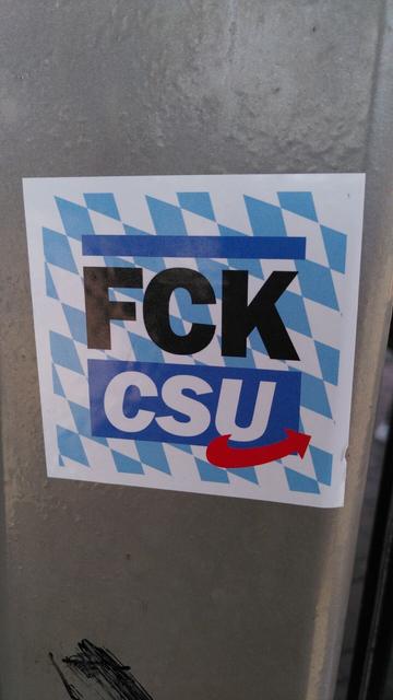 An einer Metallsäule klebt ein quadratischer Aufkleber. Als erstes fällt das bayrische Rautenmuster in blau-weiß auf. Darauf steht in Großbuchstaben "FCK CSU". Unter das CSU ist noch ein roter Pfeil gesetzt, der auf das Logo der rechtsradikalen AfD anspielt.