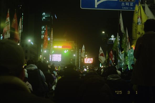 어두운 도심 속, 민주노총 조합원들이 고공투쟁 중인 고진수 관광레저산업노조 세종호텔지부장을 바라보고 있다. 무지개 깃발을 휘두르는 고진수 지부장의 모습이 어렴풋이 보인다.