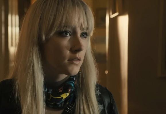 Saoirse Ronan in a close up. she has long blonde hair and is standing in a shadowy room looking slightly to her left.