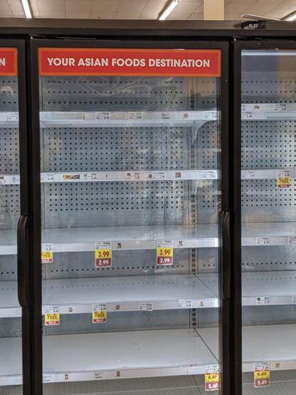 Shelves behind a glass door in a supermarket. There are a few price labels attached to the shelves, but the shelves are empty of any merchandise. A sign adhered to the top of the door reads in plain letters: "Your Asian Foods Destination". It's impossible to read the product names on the price labels. But two labels say $3.99 marked down to $2.99. Two others say $6.49 marked down to $5.49. And several labels say "Buy 1 get 1 free". The ceiling is barely visible in the photo past the shelves: a high tiled ceiling with long fluorescent lights at regular intervals.
