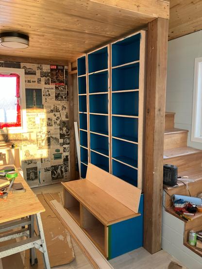 A set of shelves built in to a small wall under a loft at the cabin. A small bench is made before them. The wall behind is plastered with pages from old magazines.