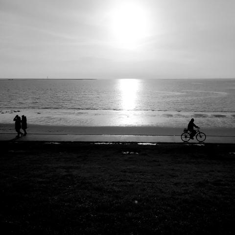 Schwarzweißfoto. Blick vom Fuße eines Deiches auf das spiegelglatte Meer, dazwischen ein Weg, links gehen zwei Fußgänger, rechts fährt ein Radfahrer. Die Personen sind nur als schwarze Silhouetten im Gegenlicht zu erkennen. Am oberen Bildrand die milchig durch den Dunst scheinende Sonne, die sich im Meer spiegelt.