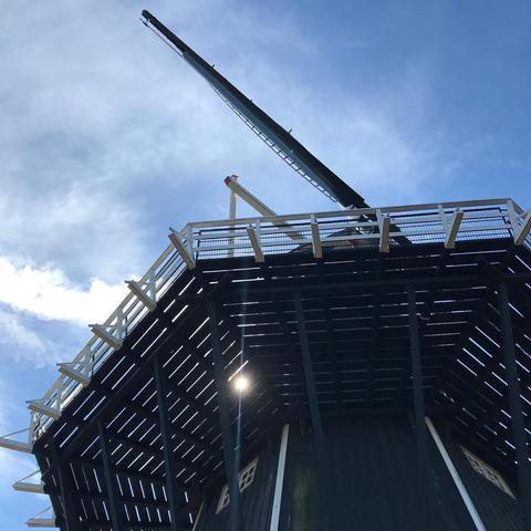 Das Bild zeigt eine Windmühle von unten aufgenommen. Der Himmel ist blau mit einigen Wolken. Die Windmühle selbst ist dunkel und hat lange, dunkle Flügel.