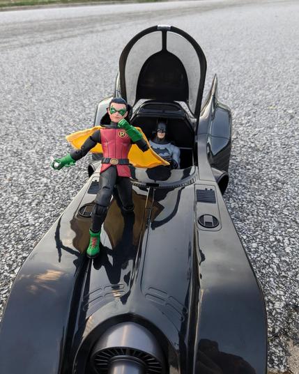 3 McFarlane toys on a paved surface: the 1989 movie Batmobile, Batman from Batman Reborn driving the Batmobile, and Damian Wayne as Robin on the hood of the Batmobile