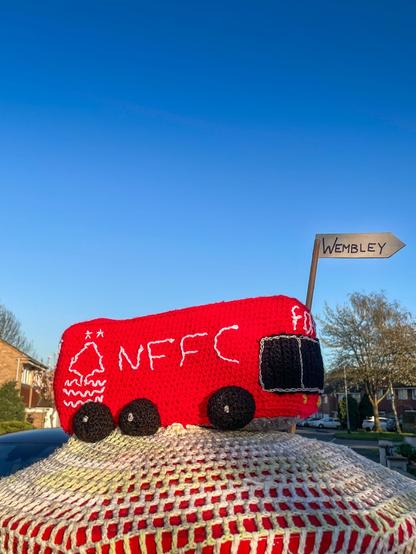 Postbox Topper.
Knitted/Crocheted red coach featuring the tricky tree and "NFFC" and a road sign pointing to Wembley.

Found on the postbox at 52.9458235, -1.2445825
