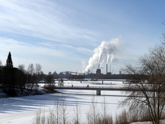 View over wintery Oulu River, with pedestrian bridges seen in distance, with pedestrians and bicyclists. The river is mostly frozen. Paper mills can be seen in far distance, spouting vapour in the air. The sky is semi-cloudy.