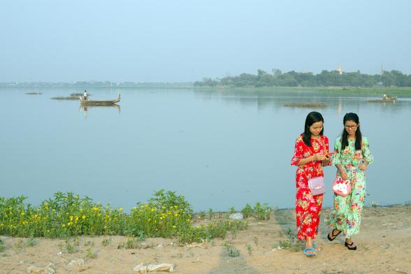 Photo shows two young ladies at the edge of a larger lake in afternoon soft sunlight just before the sunset. They are modestly dressed in matching dresses, one in red and one green basic colour. They have cute pink handbags, and are focused on their smart phone. on the lake there i a boat with a single person punting. Small islands of grass and bushes are spread out. In the background the other edge of the lakes shows areas of green and trees, and some houses and a couple of pagodas showing their roof and towers. The edge of the lake is covered in red  earth and green bushes and some yellow flowers. It is the end of a warm day, and the cloud are light blue with some mist.