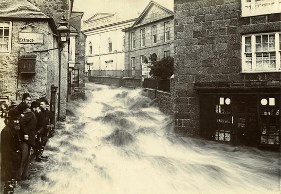 A black and white photograph of floods in St Ives, Cornwall from 1894. A powerful torrent of water is rushing down a narrow street people watch from the close to the edge. The long exposure of the camera blurs and add movement to the water.