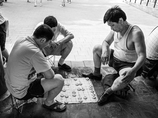 Three middle aged chinese men play chinese chess on the floor of a park in Beijing. It's summer and these men wear tank tops and t-shirts. The picture is in black and white