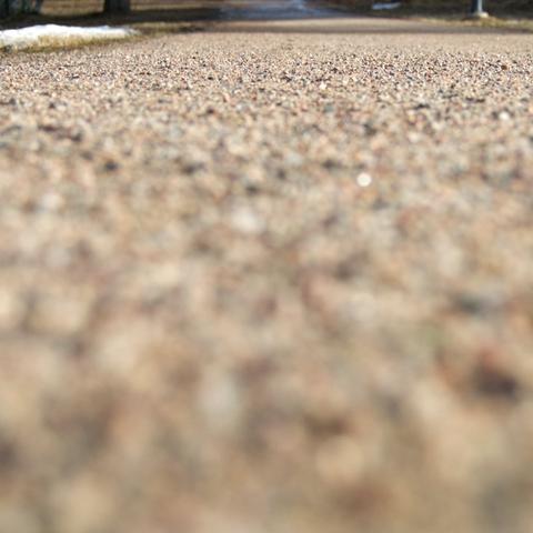 A photograph taken from ground level. Gravel and sand on top of a bicycle path, camera pointed to the distance. A stretch of gravel in near distance is in focus, the areas closer to the camera (and farther off) out of focus.
