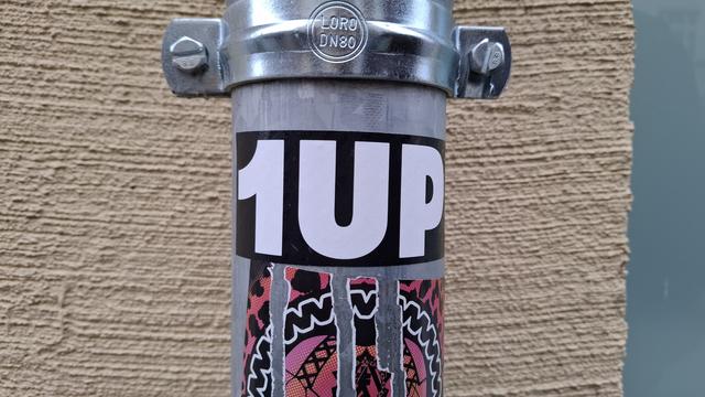 Graues Regenrohr vor einer beigen Wand, rechts eine türkisfarbene Wand angrenzend.

Auf dem Regenrohr ein großer, schwarzer Aufkleber mit "1UP" als weiße Schrift.

Darunter ein anderer bunter Aufkleber.