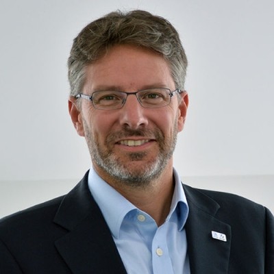 This professional headshot shows someone wearing a dark suit jacket and light blue button-down shirt against a plain light background. The person has short greyish hair, glasses, and a neatly trimmed grey beard. They are smiling warmly at the camera. A small organizational pin or badge is visible on their lapel.