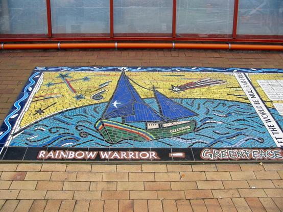 Ein buntes, rechteckiges Mosaik am Rand eines Gehweges, das das Greenpeace-Schiff Rainbow Warrior auf dem Ozean zeigt. Am Rand steht „Let the world be nuclear free.“