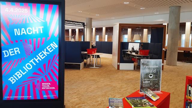 Innenansicht der SLUB Dresden. Ein Monitor mit der Aufschrift Nacht der Bibliotheken mit roten und blauen Strahlen. Ein Tisch mit Büchern zum Thema Wald.
Foto: © SLUB Dresden, Annemarie Grohmann