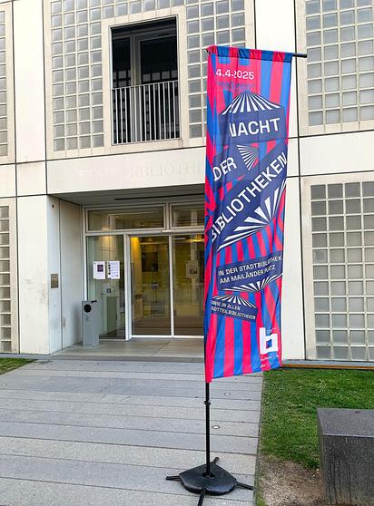 Eine Fahne vor einem Gebäude mit der Aufschrift:
 4.4.2025
Nacht der Bibliotheken.
Im Hintergrund sieht man den Eingang eines Bibliotheksgebäudes.
Foto: Stadtbibliothek Stuttgart
