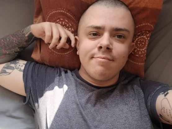 A white trans masc wearing a grey t shirt. He's lying in bed and has a shaved head. His right arm is raised. You can see tattoos on both arms