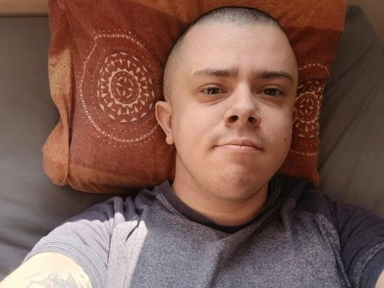 A white trans masc wearing a grey t shirt. He's lying in bed and has a shaved head