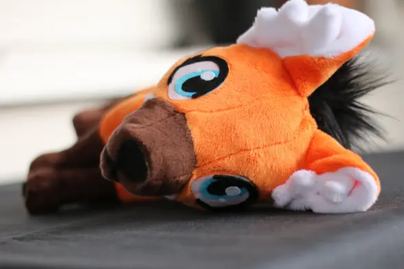 An orange, brown and white maned wolf plushie lying on the side. It has a fluffy black mane on the back and really large eyes.