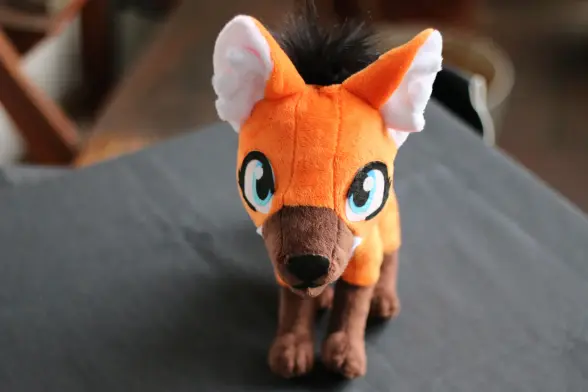 An orange maned wolf plushie placed on grey table cloth to look towards the camera in a rather cute way.