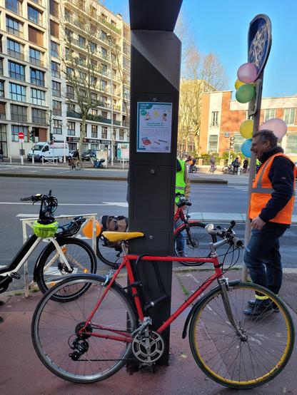 Vélo vitrage rouge posé contre un poteau. Bénévoles en gilets jaune et orange.