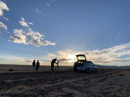 Tres personas, dos mujeres y un hombre, están de pie en un paisaje desértico plano bajo un cielo azul claro y soleado.  El hombre está usando un trípode de cámara, sugiriendo que están grabando o fotografiando.  Un coche plateado con la cajuela abierta está estacionado cerca. El sol está bajando, creando una iluminación suave y dorada en el horizonte.  La escena general transmite una sensación de tranquilidad y espacio abierto.