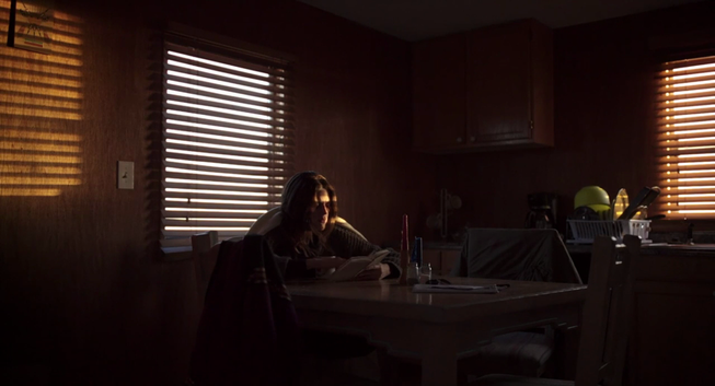 Una joven con el cabello largo y oscuro está sentada a una mesa en una cocina oscura y poco iluminada. Está leyendo un libro y la luz del sol entra por una ventana con persianas de madera, creando sombras en la pared. La cocina tiene un ambiente rústico y la mesa está parcialmente iluminada por la luz del sol.