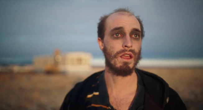 Un hombre joven, con la cabeza casi sin pelo y una barba corta, mira hacia arriba con una expresión de sorpresa o miedo. Tiene los ojos maquillados con sombra oscura, dando una apariencia ligeramente fantasmal. Viste una chaqueta oscura. El fondo está desenfocado, mostrando un paisaje desértico con una caravana en la distancia bajo un cielo crepuscular.