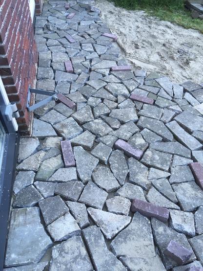A cobblestone pathway made up of irregularly shaped gray stones, interspersed with a few reddish-purple bricks, alongside a patch of sand and grass. The pathway runs along the side of a building.