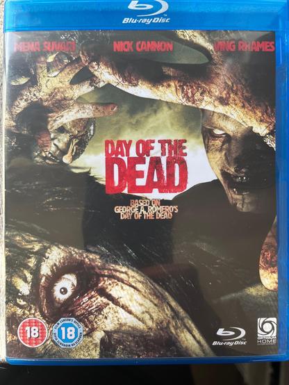 The image shows a Blu-ray disc case for the movie "Day of the Dead." The case is predominantly blue with a clear plastic cover. The title "Day of the Dead" is prominently displayed in large, bold red letters in the center. Below the title, it states "Based on George A. Romero's Day of the Dead." The names of the actors, Mena Suvari, Nick Cannon, and Ving Rhames, are listed at the top in red text. The cover features a graphic of a zombie with a close-up of its face and hands, conveying a sense of horror and suspense. The background is dark and gritty, enhancing the eerie atmosphere. The Blu-ray logo is visible at the top, and the Optimum Home Entertainment logo is at the bottom right. The age rating is 18, as indicated by the red and blue stickers on the bottom left.