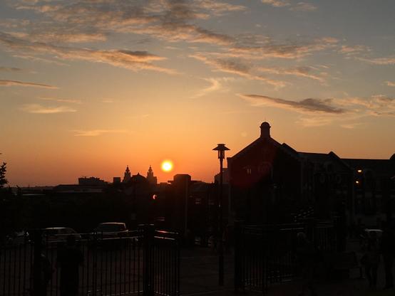The bottom of the image shows a dark silhouette of Liverpool, while the top shows the sky, featuring a blue to orange gradient (top to bottom). The sun is in middle, close to setting behind the city silhouette. Right next to the sun, one can recognise the Royal Liver Building.