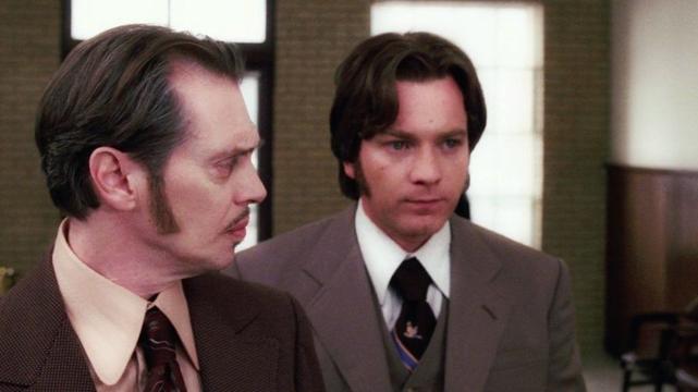 Two men in suits, with old-fashioned haircuts with long sideburns, stand inside a building.