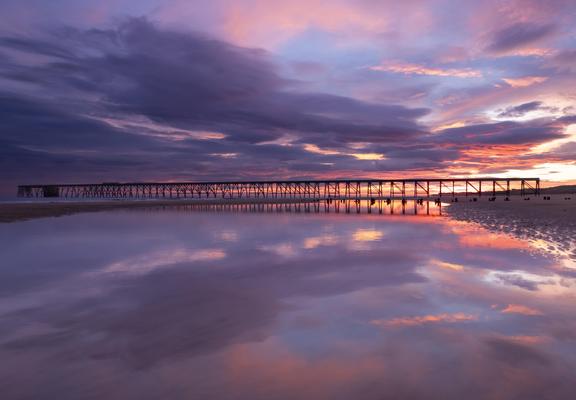 A photo of a old wooden industrial pier during the sunrise blue hour. The sky is a kaleidoscope of orange, pink, blue, red and orange above the pier. The sky is partially reflected in a pool of sea water on the beach. 

Whilst I was taking this my feet were slowly sinking in the sand which meant ice cold water was rushing into my walking boots.