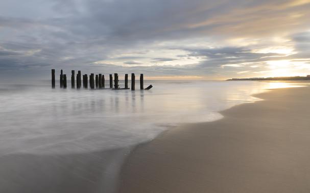A long exposure photo of remnants of a pier that are standing upright as waves lap around them. The sun can be seen rising to the right of the frame casting a golden light on the scene as waves break in the foreground.