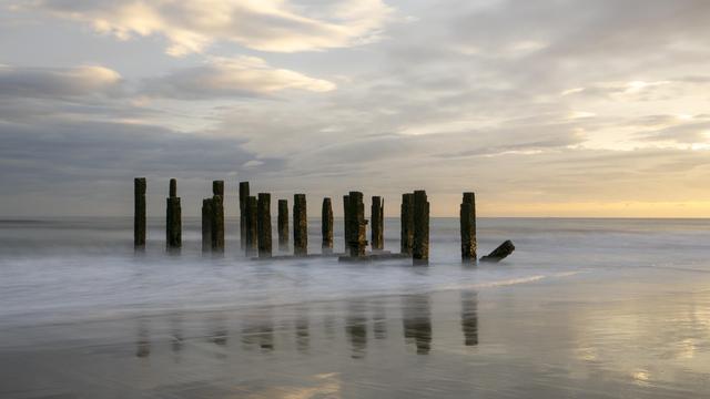 A long exposure photo of remnants of a pier that are standing upright as waves lap around them. The sun is rising to the right of the frame casting a golden light on the scene.