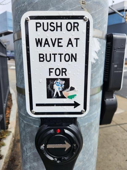 Metal pole holding crosswalk button with sign that says: Push or Wave at Button for ------>

Stickers or scratches obscure some of the picture of a pedestrian.