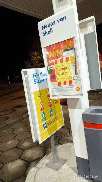 Foto von einer Ankündigungstafel auf einer Shelltankstelle. Der Infobereich ist mit einem rot-gelben Zettel mit Shelllogo in der rechten unteren Ecke überklebt, auf dem steht: "Kaputtes System." Klein darüber: "Wir sorgen für ein" und darunter: "Das tut uns leid."