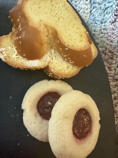 Photo of two slices of challah bread, and two "thumbprint" jam cookies (called bird's nest cookies by the bakery, which is Gunn's), on a black surface with a softly multicolored afghan in the background.