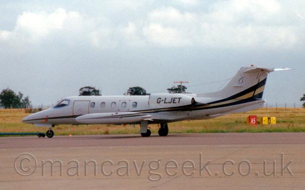Slightly grainy side view of a white, twin engined BizJet, parked facing to the left.
It appears to have a long, green towbar attached to it's nosewheel.
The plane is mostly white, with a black stripe with a gold stripe down the middle, running along the body, getting wider as it moves to the rear, sweeping up into the lower reaches of the tail.
Grey concrete apron fills the foreground, with grass visible below the plane leading up to a chain-link fence and scattered trees in the distance, under flat grey skies.